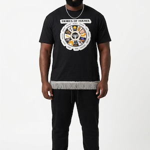 May include: Black t-shirt with a circular graphic displaying the words "Tribes of Israel" and various symbols. The shirt is worn with black sweatpants and white trainers. A silver chain is worn around the neck. The outfit includes a decorative fringe.