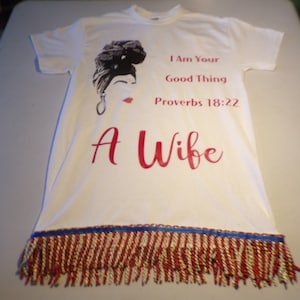 May include: White t-shirt with a black and white illustration of a woman with a headscarf. The text "I Am Your Good Thing Proverbs 18:22 A Wife" is printed in red on the shirt. The bottom of the shirt has a red, white, and blue fringe.