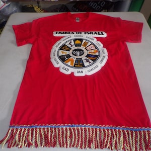 May include: Red t-shirt with a white circle design featuring the "Tribes of Israel" text and symbols. The shirt has a fringe trim with red, blue, and gold threads.