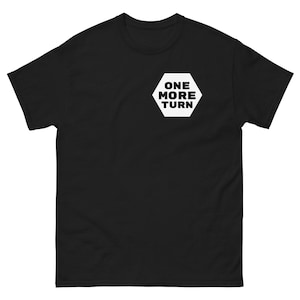 Op de afbeelding: Zwart T-shirt met een witte zeshoekige patch waarop in hoofdletters "ONE MORE TURN" staat.