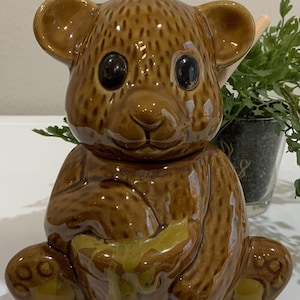 May include: A brown ceramic bear-shaped honey pot with a glossy finish. The bear is sitting, holding a honey-filled bowl. It has large black eyes and small ears. The honey pot is a decorative kitchen item.