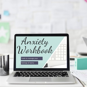 May include: A laptop displaying the words "Anxiety Workbook" with the subtitle "Tools to manage your feelings of worry." A silver mug, a small teal box, and a pen are on the desk. A second laptop is visible in the background.