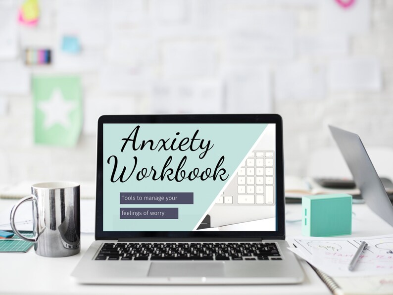 May include: A laptop displaying the words "Anxiety Workbook" with the subtitle "Tools to manage your feelings of worry." A silver mug, a small teal box, and a pen are on the desk. A second laptop is visible in the background.