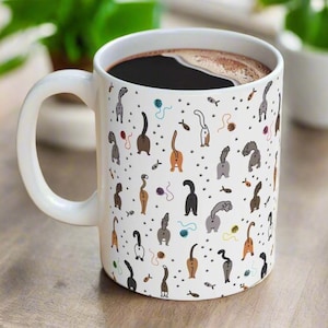 May include: White ceramic coffee mug with a pattern of colorful cats' rear ends. The mug is filled with coffee.