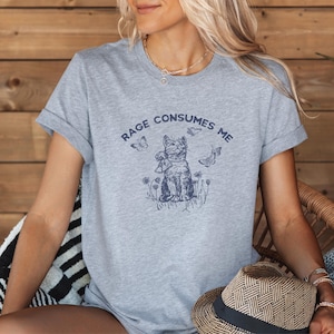 以下が含まれることがあります： 袖をまくったヘザーグレーのTシャツ。紺色の猫のグラフィックが描かれており、蝶と花に囲まれています。「RAGE CONSUMES ME」の文字が猫のイラストの上にアーチ状に配置されています。麦わら帽子が画像の下部に表示されています。