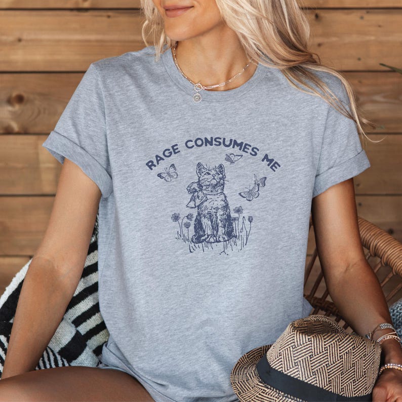 以下が含まれることがあります： 袖をまくったヘザーグレーのTシャツ。紺色の猫のグラフィックが描かれており、蝶と花に囲まれています。「RAGE CONSUMES ME」の文字が猫のイラストの上にアーチ状に配置されています。麦わら帽子が画像の下部に表示されています。