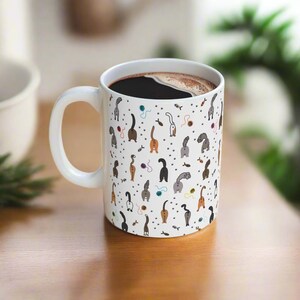 May include: White ceramic coffee mug with a black handle and a pattern of cats' rear ends in various colors and poses. The mug is filled with coffee.