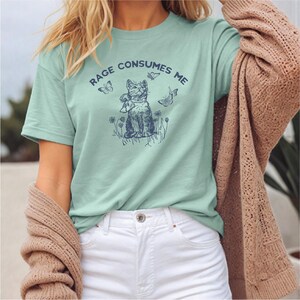 以下が含まれることがあります： 「RAGE CONSUMES ME」と紺色の文字がプリントされた、薄緑色のTシャツ。蝶と花に囲まれた猫の紺色のイラストが特徴です。柔らかい素材で作られたTシャツです。