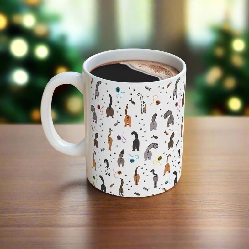 May include: White ceramic coffee mug with a black handle. The mug is covered in a pattern of cartoon cats' rear ends in various colors and poses. The mug is filled with black coffee.