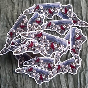 Puede incluir: Un conjunto de pegatinas con la forma del estado de Carolina del Norte. Cada pegatina presenta un p&aacute;jaro cardenal rojo posado en una rama floreciente, con una cordillera al fondo. Las pegatinas tienen bordes blancos.