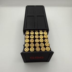 May include: A black container filled with brass-colored bullets. The container has a hinged lid and the number "556" in red lettering on the front. The bullets are arranged neatly inside the container.