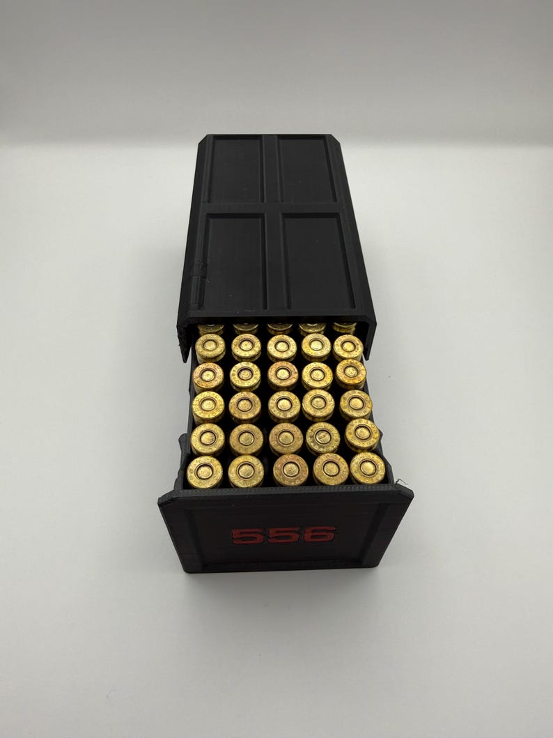 May include: A black container filled with brass-colored bullets. The container has a hinged lid and the number "556" in red lettering on the front. The bullets are arranged neatly inside the container.