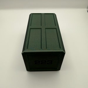 May include: A dark green rectangular box with a textured surface. The top has a recessed cross pattern. The number "223" is printed on the front of the box. The box is sitting on a white surface.