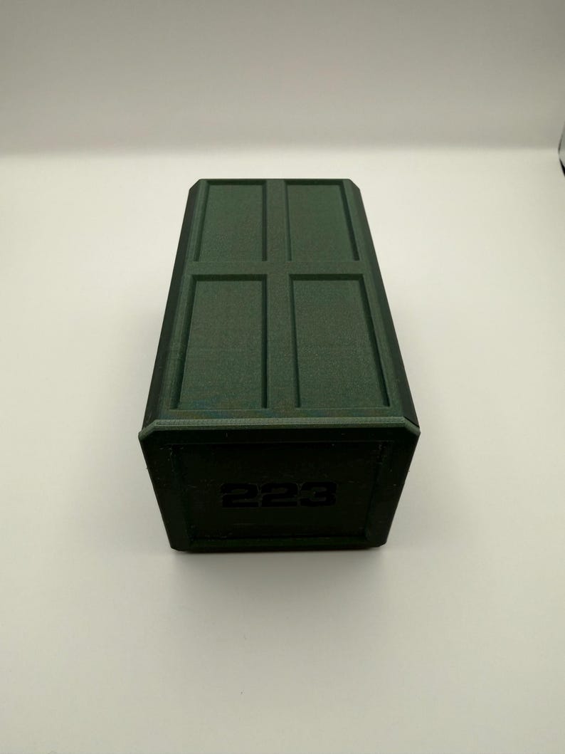 May include: A dark green rectangular box with a textured surface. The top has a recessed cross pattern. The number "223" is printed on the front of the box. The box is sitting on a white surface.