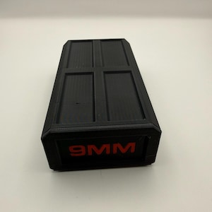 May include: A black rectangular container with a textured surface and a red "9MM" label. The top of the container is divided into four rectangular sections.