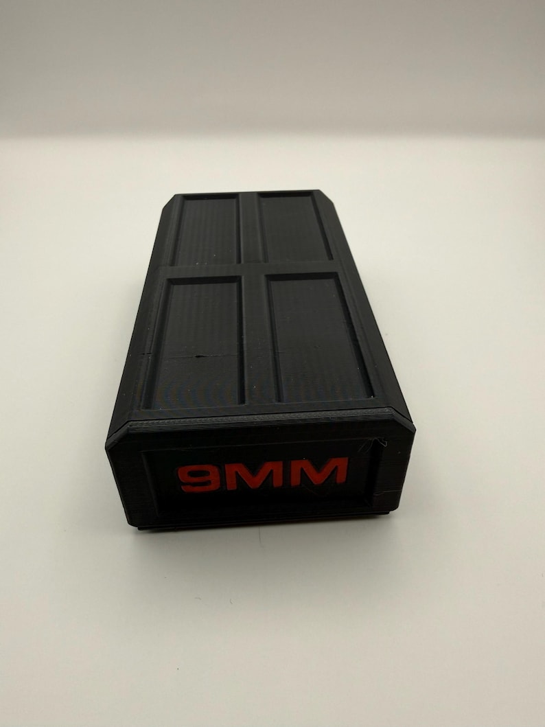 May include: A black rectangular container with a textured surface and a red "9MM" label. The top of the container is divided into four rectangular sections.