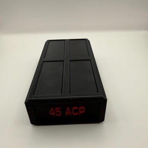 May include: A black rectangular container with a grid pattern on the top surface. The text "45 ACP" is printed in red on the side of the container. The container is likely used for storing ammunition.