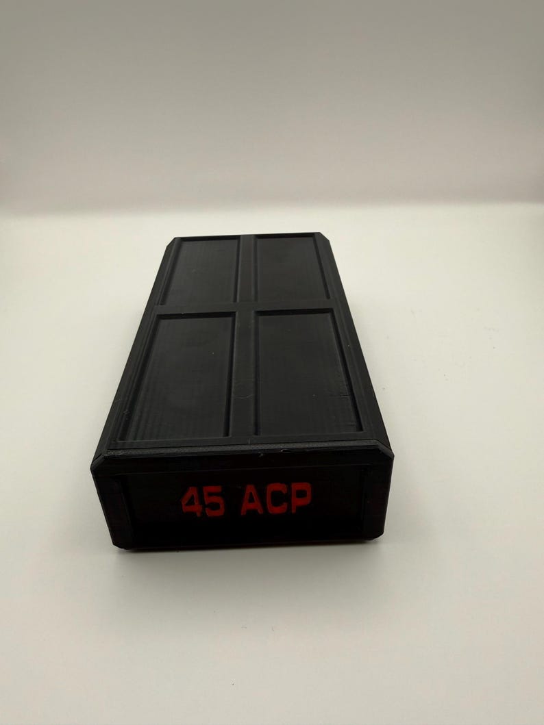 May include: A black rectangular container with a grid pattern on the top surface. The text "45 ACP" is printed in red on the side of the container. The container is likely used for storing ammunition.