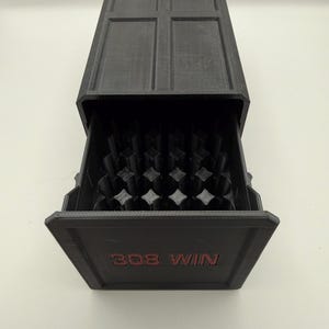 May include: A black rectangular storage box with a sliding drawer. The drawer is open, revealing a grid of objects. The text "308 WIN" is printed in red on the front of the drawer. The top of the box has a cross-shaped design.