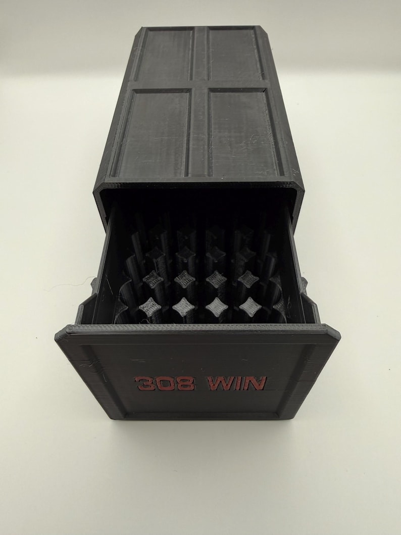 May include: A black rectangular storage box with a sliding drawer. The drawer is open, revealing a grid of objects. The text "308 WIN" is printed in red on the front of the drawer. The top of the box has a cross-shaped design.