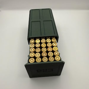May include: A dark green ammunition box with a hinged lid, filled with brass-colored bullets. The box is labeled with the number "223" on the front. The bullets are neatly arranged inside the box.