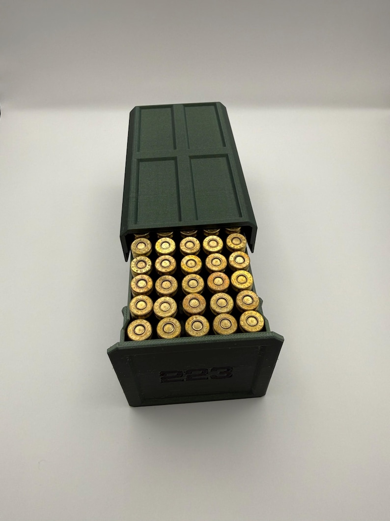 May include: A dark green ammunition box with a hinged lid, filled with brass-colored bullets. The box is labeled with the number "223" on the front. The bullets are neatly arranged inside the box.