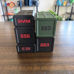 May include: Four stacked black and green boxes with red text. The boxes are labeled with the following text: 9MM, 556, 40 S&W, and 223. The boxes are sitting on a wooden surface.
