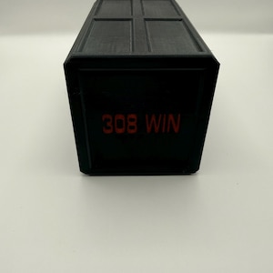 May include: A black rectangular box with a hinged lid. The text "308 WIN" is printed in red on the front of the box. The box has a matte finish and appears to be made of plastic.