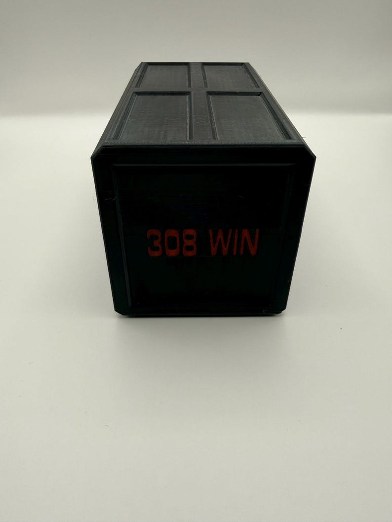 May include: A black rectangular box with a hinged lid. The text "308 WIN" is printed in red on the front of the box. The box has a matte finish and appears to be made of plastic.