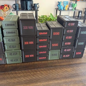 May include: A collection of stacked, rectangular ammunition boxes. The boxes are in various colors, including black and olive green. Each box is labeled with red or white text indicating the ammunition type, such as "556", "9MM", "45 ACP", and "308 WIN".