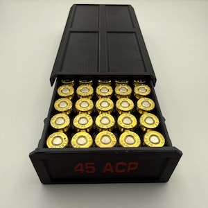 May include: A black box with a hinged lid, filled with gold-colored bullets. The box has the text "45 ACP" in red on the front. The bullets are arranged neatly inside the box.