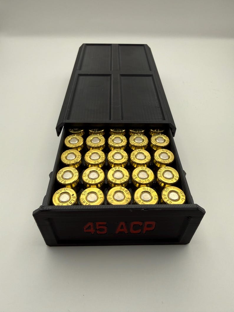 May include: A black box with a hinged lid, filled with gold-colored bullets. The box has the text "45 ACP" in red on the front. The bullets are arranged neatly inside the box.