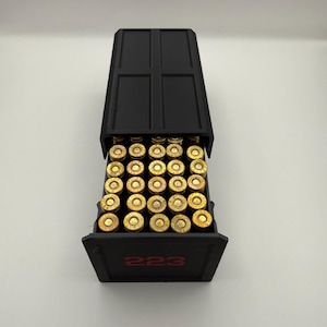 May include: A black ammunition box with a hinged lid, filled with numerous brass-colored cartridges. The number "223" is printed in red on the front of the box. The box is open, revealing the contents.