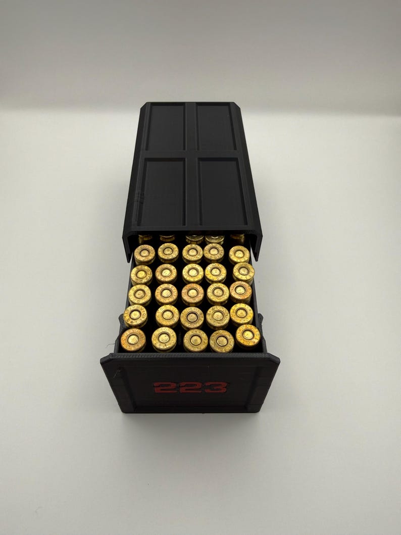 May include: A black ammunition box with a hinged lid, filled with numerous brass-colored cartridges. The number "223" is printed in red on the front of the box. The box is open, revealing the contents.