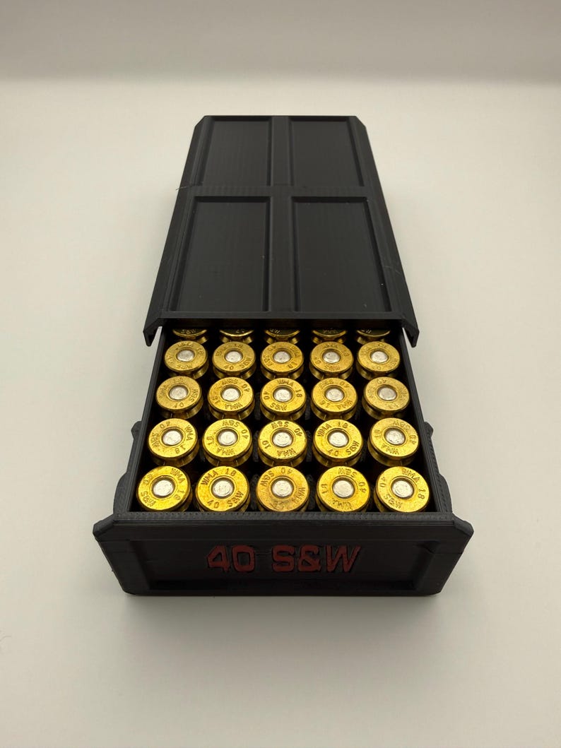 May include: Black 3D-printed ammunition box with a hinged lid, open to reveal rows of gold-colored bullets. The box is labeled "40 S&W" in red lettering, indicating the ammunition type. The bullets are arranged neatly inside.