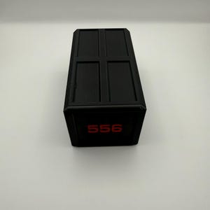 May include: A black rectangular box with a grid pattern on the top. The number "556" is displayed in red on the front of the box. The box is sitting on a white surface.