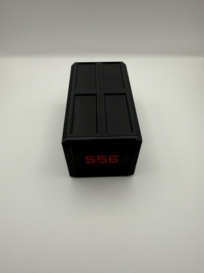 May include: A black rectangular box with a grid pattern on the top. The number "556" is displayed in red on the front of the box. The box is sitting on a white surface.
