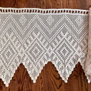 May include: An off-white crochet valance featuring a repeating diamond pattern and a scalloped bottom edge. The top section includes a row of open circular loops for hanging, with the intricate needlework creating a textured, geometric design.
