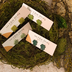 May include: Three pairs of green shamrock-shaped earrings on white cards with the text "rose & fern artistry handcrafted jewellery".