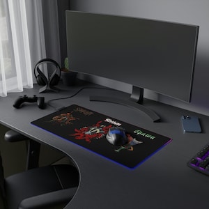 May include: A black gaming mousepad with a colorful graphic of a comic book character. The mousepad has a blue and purple LED light around the edge. The mousepad is on a black desk with a computer monitor, a gaming controller, and a smartphone.