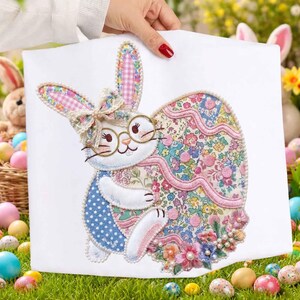 Pode incluir: Uma peça decorativa branca com tema de Páscoa, apresentando um coelho com óculos e segurando um ovo floral. O coelho tem detalhes em rosa e azul, com um laço e detalhes florais. O ovo é adornado com padrões florais coloridos e uma borda rosa.