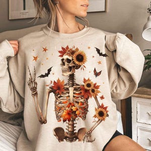 May include: A beige sweatshirt with a skeleton wearing a crown of fall leaves and flowers. The skeleton is holding a peace sign with its left hand. The sweatshirt has a relaxed fit and long sleeves.