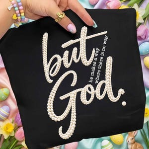 以下が含まれることがあります： 黒いTシャツに「but God」の文字が真珠のようなデザインで施されています。「he makes a way where there is no way」というフレーズも表示されています。Tシャツは、金の指輪とカラフルなブレスレットをつけた人によって持たれています。