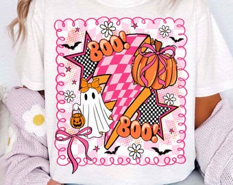 Puede incluir: Camiseta blanca con un diseño de Halloween. El diseño incluye un rayo rosa y naranja, una calabaza con un lazo, un fantasma y la palabra "Boo!" en naranja. El diseño está enmarcado con un borde rosa.