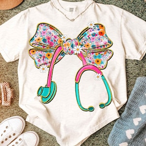 Peut inclure: T-shirt blanc avec un motif de n&oelig;ud floral et de st&eacute;thoscope. Le n&oelig;ud est orn&eacute; de fleurs color&eacute;es et bord&eacute; d'or. Le st&eacute;thoscope est rose et turquoise, avec des accents de marguerites. Un collier est visible au d&eacute;collet&eacute;.