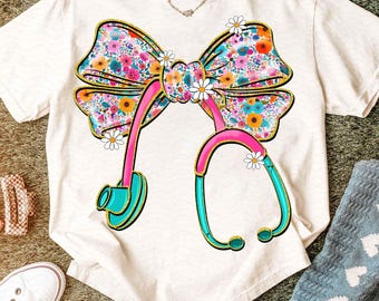 Pode incluir: Camiseta branca com um design de laço floral e estetoscópio. O laço é adornado com flores coloridas e contornado em dourado. O estetoscópio é rosa e turquesa, com pequenos detalhes de margaridas. Um colar é visível no decote.