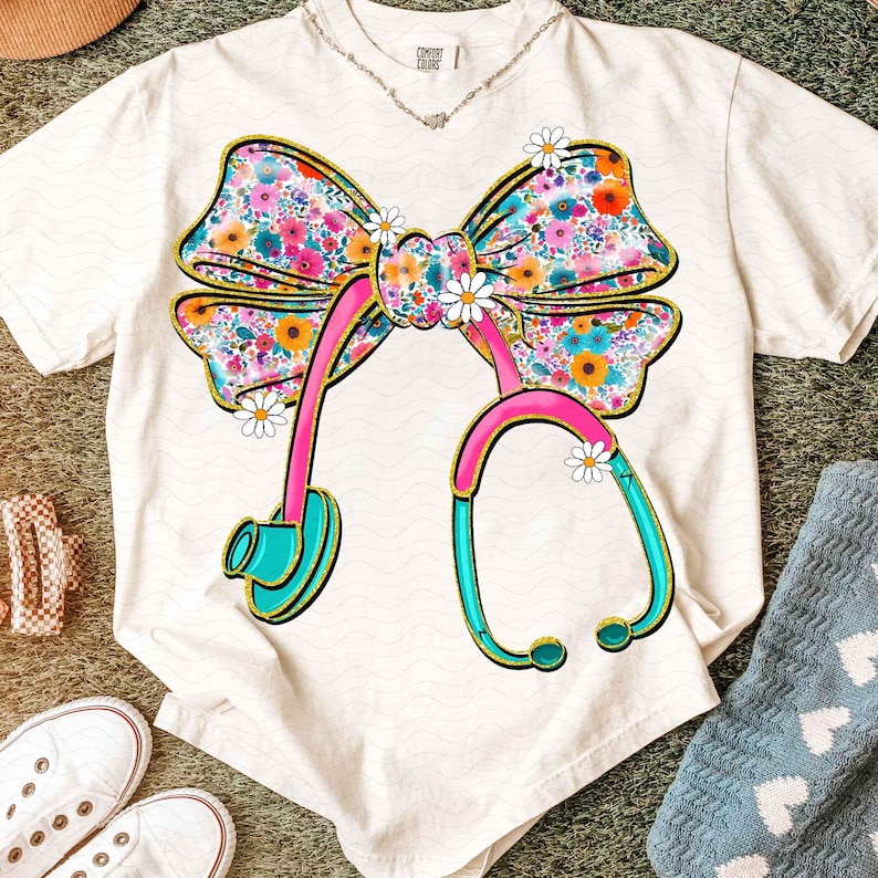 Peut inclure: T-shirt blanc avec un motif de n&oelig;ud floral et de st&eacute;thoscope. Le n&oelig;ud est orn&eacute; de fleurs color&eacute;es et bord&eacute; d'or. Le st&eacute;thoscope est rose et turquoise, avec des accents de marguerites. Un collier est visible au d&eacute;collet&eacute;.