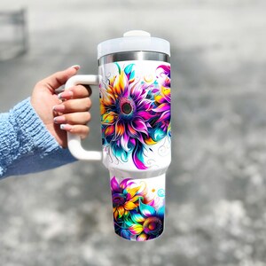 Pode incluir: Caneca de viagem branca com uma al&ccedil;a grande, com desenhos vibrantes e coloridos de girass&oacute;is. Os girass&oacute;is t&ecirc;m tons de amarelo, rosa, roxo e azul-petr&oacute;leo. A caneca tem uma tampa branca e um interior de a&ccedil;o inoxid&aacute;vel.