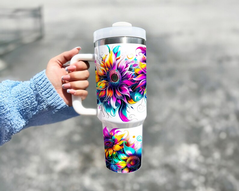 Pode incluir: Caneca de viagem branca com uma al&ccedil;a grande, com desenhos vibrantes e coloridos de girass&oacute;is. Os girass&oacute;is t&ecirc;m tons de amarelo, rosa, roxo e azul-petr&oacute;leo. A caneca tem uma tampa branca e um interior de a&ccedil;o inoxid&aacute;vel.