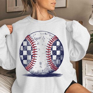 Puede incluir: Sudadera blanca con un gr&aacute;fico de una pelota de b&eacute;isbol con un patr&oacute;n a cuadros. La pelota de b&eacute;isbol est&aacute; dibujada en un estilo vintage.